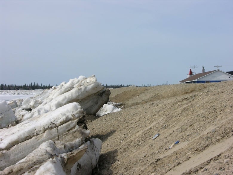 Large chunks of ice creep up the side of an earthen dike with a rooftop in the distance  