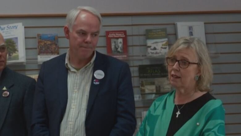 A man in a suit stands besides a woman with glasses wearing green.