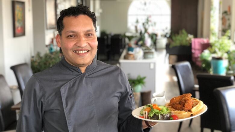 A man in a chef's jacket holding a plate of chicken and waffles in a restaurant.