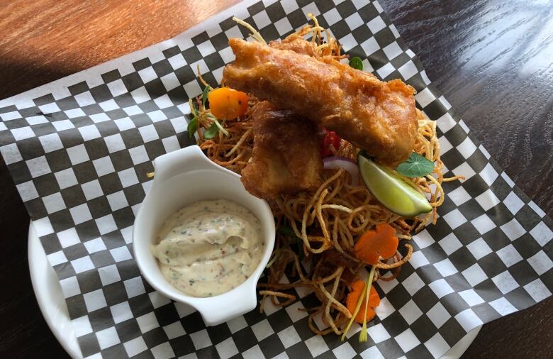 Two pieces of battered, fried fish sit atop a pile of crispy noodles next to a creamy sauce.