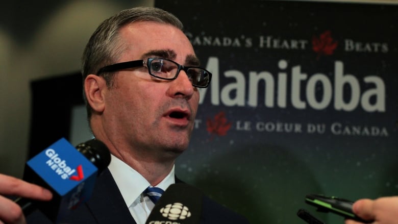 A man wearing a black suit and blue and white tie speaks in front of reporters holding microphones.