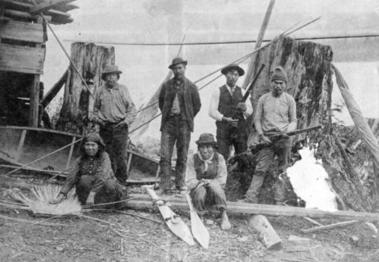 Six people sit and stand with rifles, weavings, and paddles near their community of Kitsilano Indian Reserve no. 6, also known as the ancient Skwxwu7mesh (Squamish) village Senw.