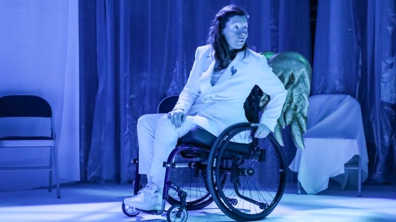 A woman sits in a wheelvhair on stage. She is wearing a pair of angel wings and is dressed in all white. She is awash in a blue light.