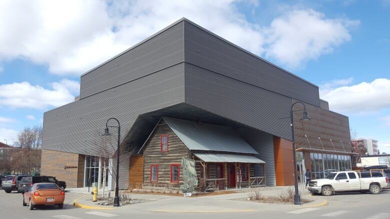 A small wooden cabin  is nestled in the outer corner of a large grey-and-brown modern building. 