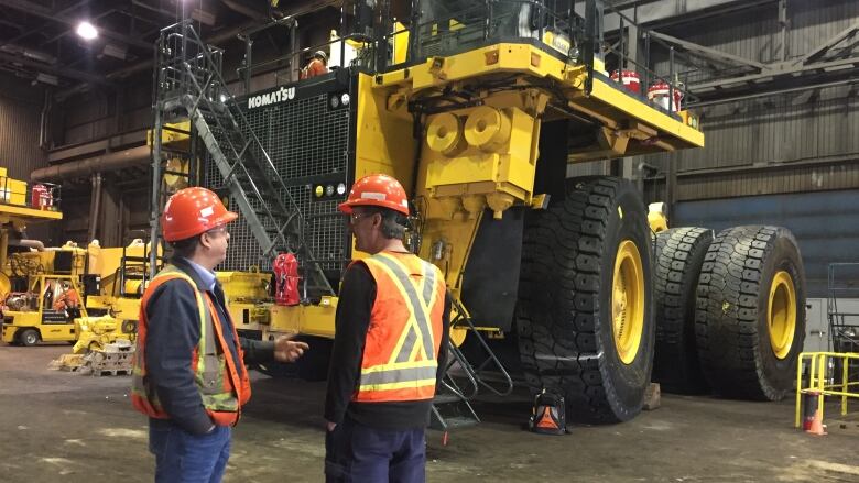 Two men in safety vest stand in front of a large truck with tires as big as the men. 