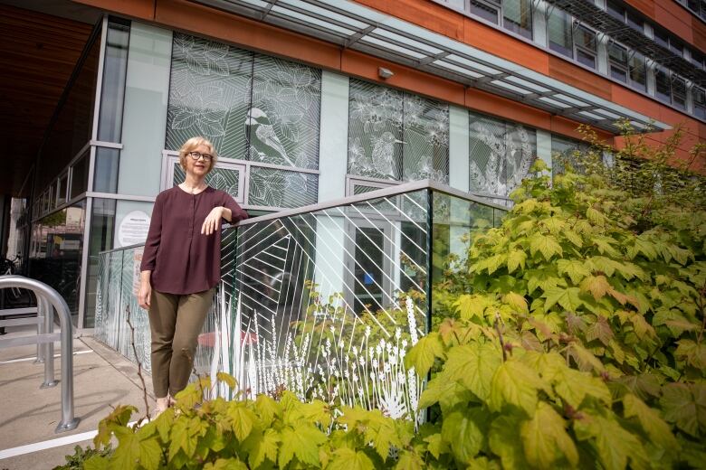 A woman wearing a purple top leans against a glass railing which has an elaborate white design on it, surrounded by green leaves.