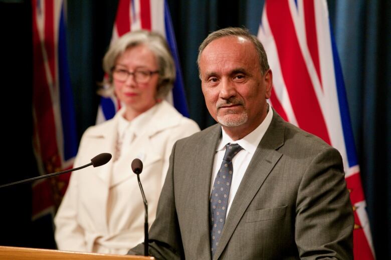 An older South Asian man delivers a news conference in front of B.C. flags, while a white woman with short hair stands behind him.