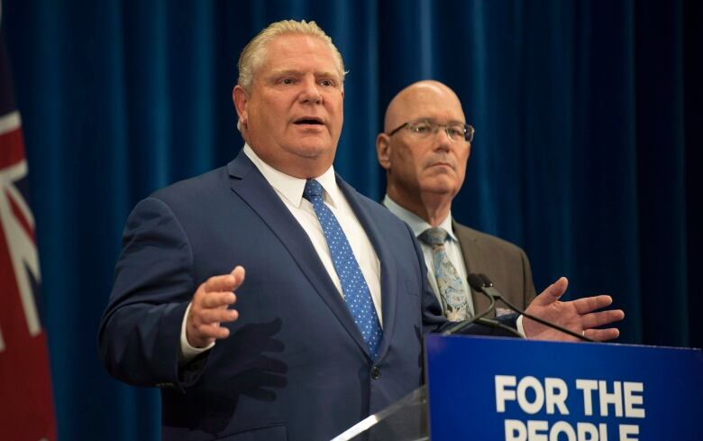 Two men speak at a press conference behind a podium sign that reads 