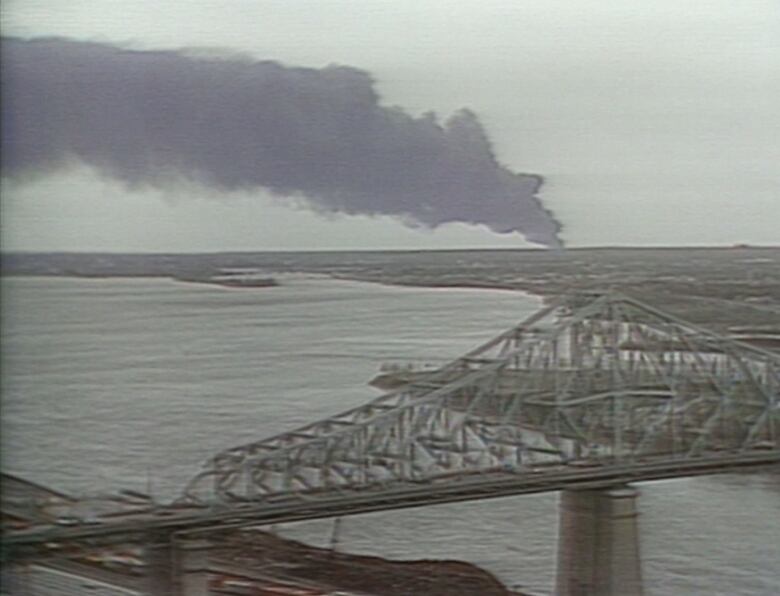 Plume of black smoke on horizon with a bridge and river in foreground