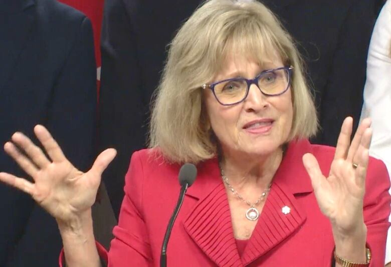 A woman in a red jacket and glasses raises her hands as she speaks.