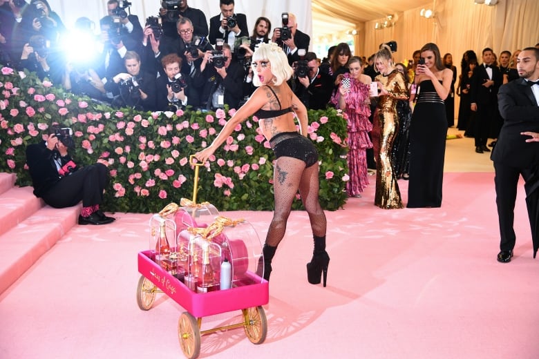 A woman in a black bikini poses  on a  pink  carpet holding a wagon