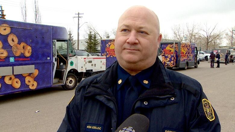 A white man wears a firefighter's uniform and speaks in front of food trucks.