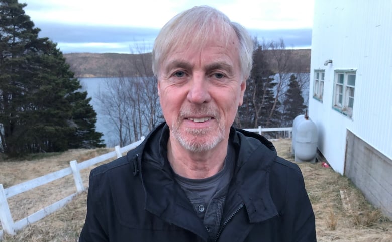 a man with white hair and a grey beard, standing outdoors wearing a black coat.