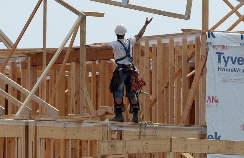 A man standing on the frame of a new home under construction, reaching his hand upwards.