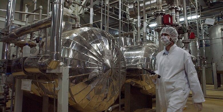 A person, head to toe in white protective clothing, walks through a room filled with a complicated array of pipes and equipment. 