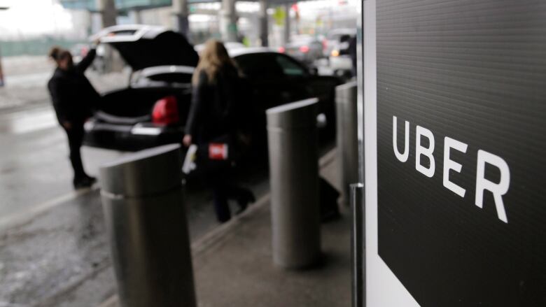 An Uber sign is in the foreground of the photo, in the background someone approaches a black vehicle while someone else loads it's trunk. 