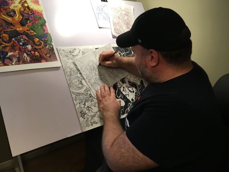 A man wearing a black tee-shirt and a black baseball hat traces a comic book drawing on an easel. His back is to the camera.