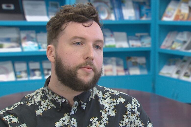 A bearded man wearing a black shirt with white flowers sits in front of an office table and bookcases holding brochures and pamphlets 