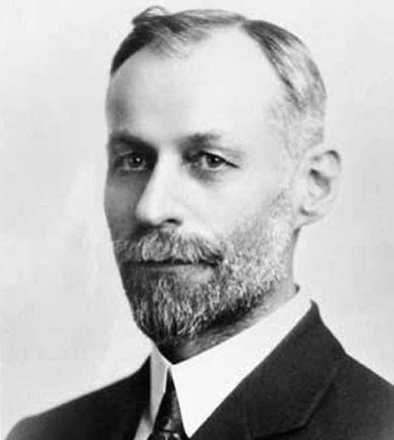 A black-and-white photo shows a man with greying hair and beard in a suit.