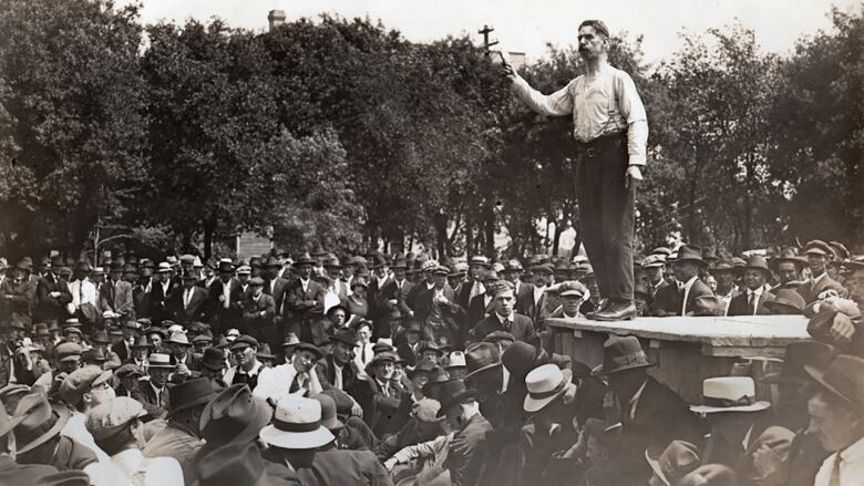 A black-and-white photo shows a crowd around a platform where a man stands, one arm up with his index finger pointing upward.