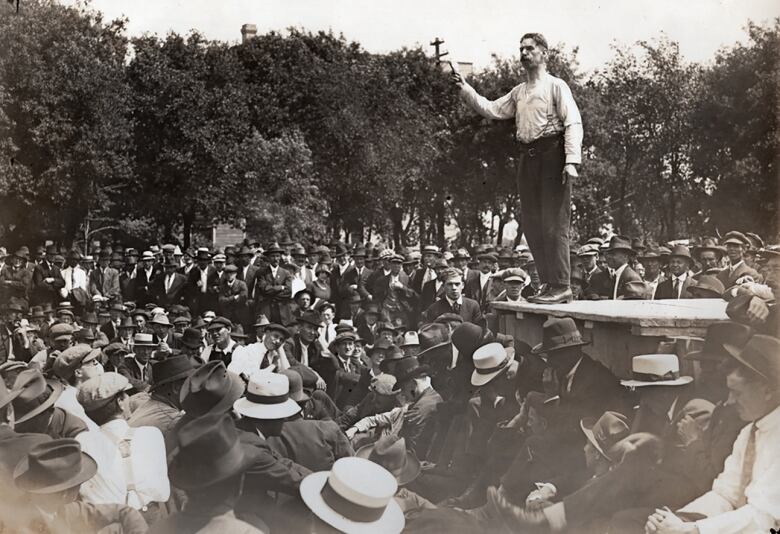 A black-and-white photo shows a crowd around a platform where a man stands, one arm up with his index finger pointing upward.