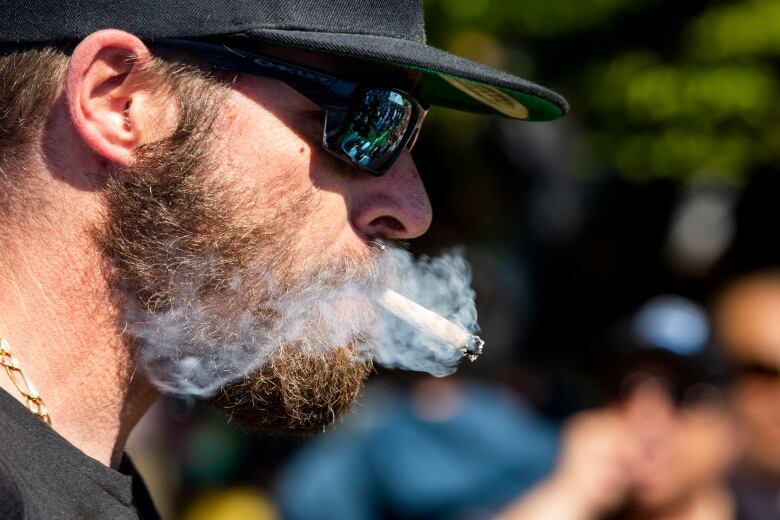 A man in a baseball cap and sunglasses shown in profile blows out a large cloud of smoke from the joint he is smoking.