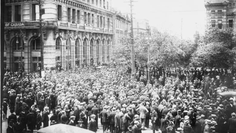 A black-and-white photo shows a big crowd of people, mostly men in suits wearing hats, standing on a street.