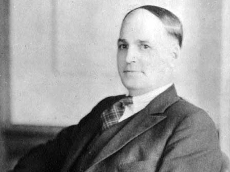 A black-and-white photo shows a man with slicked-down hair in a suit.