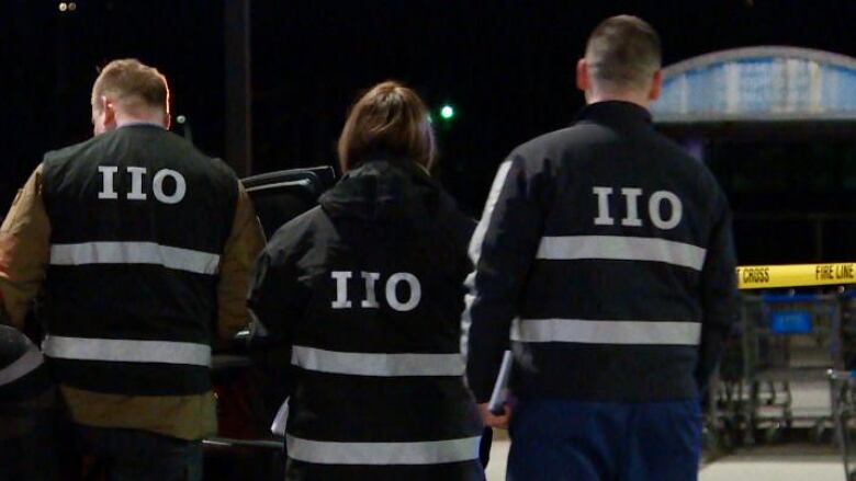 Two men stand on either side of a women and all have their back to the camera. It is dark out, but the back of their black jackets have reflector tape that spells out IIO. In front of them, is yellow police tape about waist high that says police line do not cross.