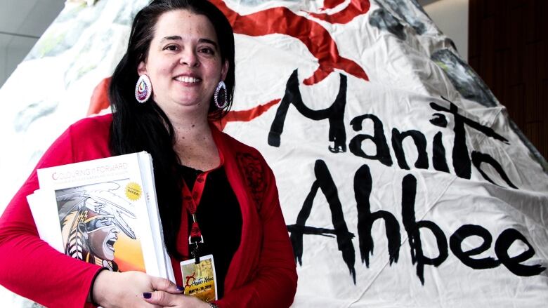 A woman with dark hair and red earrings wears a red sweater and smiles in front of a teepee