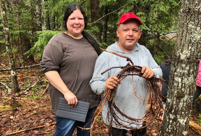 An Indigenous woman smiles standing next to an Indigenous man holding roots in a forest.