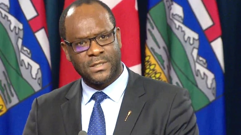 A man wearing glasses and a suit stands in front of Alberta and Canada flags.