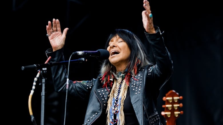 A woman wearing a leather jacket, a beaded necklace and feathers in her hair sings into a microphone.