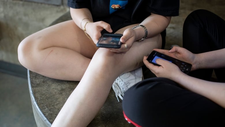 High school students, their faces unseen, sit cross-legged on concrete steps while scrolling on cellphones