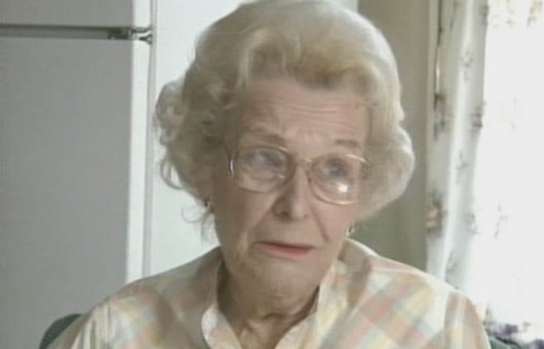 An older woman with light hair and glasses is sitting and speaking.