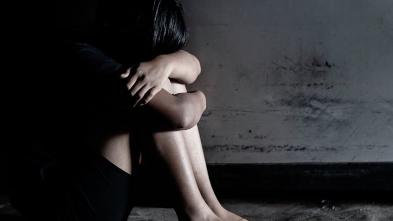 A young girl is shown sitting, holding her knees up to and covering her face.