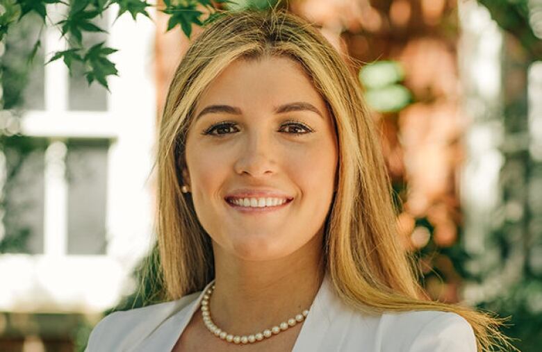 A woman with blonde hair and a pearl necklace smiles outside.