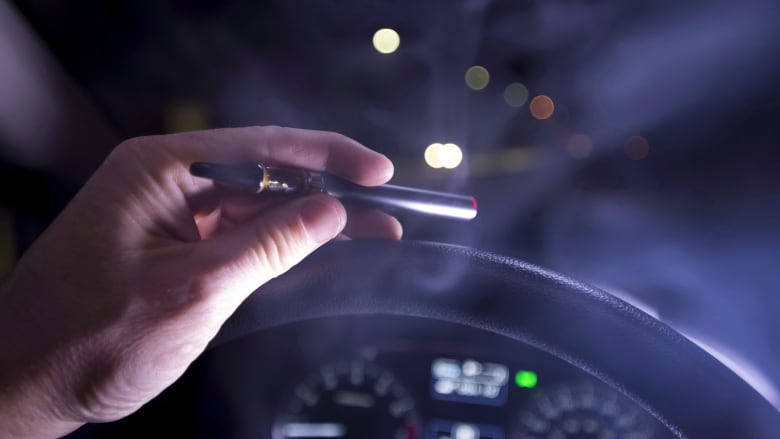 A hand holding a cannabis vaporizer while gripping a steering wheel.