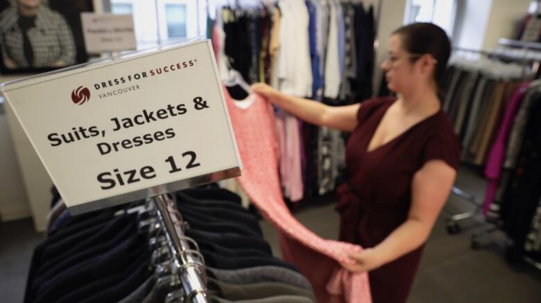 A sign on a clothing rack says Suits, Jackets and Dresses, size 12, while in the background a woman looks at the clothes.