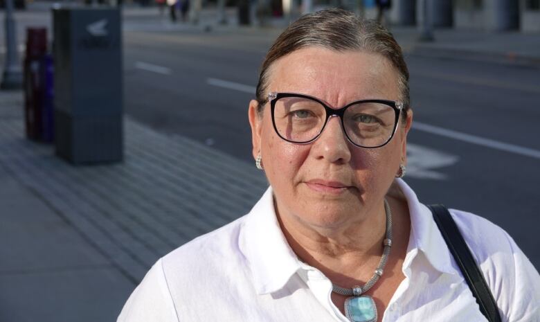 A woman wearing glasses and a white shirt stands outside on a downtown street.