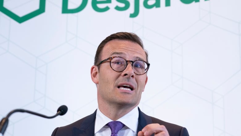 Guy Cormier has dark hair, round glasses and is wearing a suit with a blue tie. He is speaking into a small microphone and behind him it reads: Desjardins.