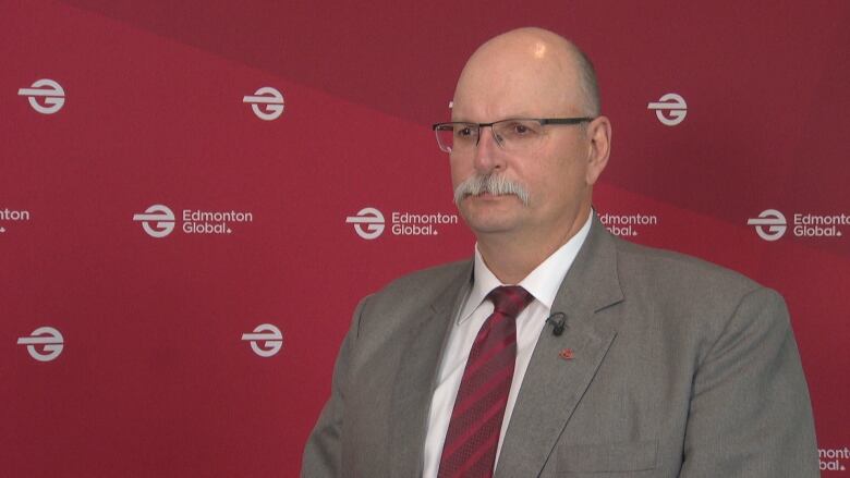 A bald man wearing glasses stands in front of a red backdrop that says 