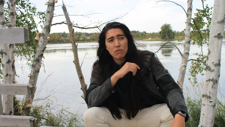 A man with long hair sits on a bench in front of a body of water with his elbow on his knee.