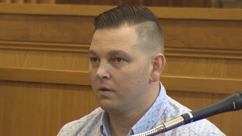 A man is wearing a dress shirt and is sitting in a wooden prisoner's box. His hair is gelled back on top, with the sides closely cropped.