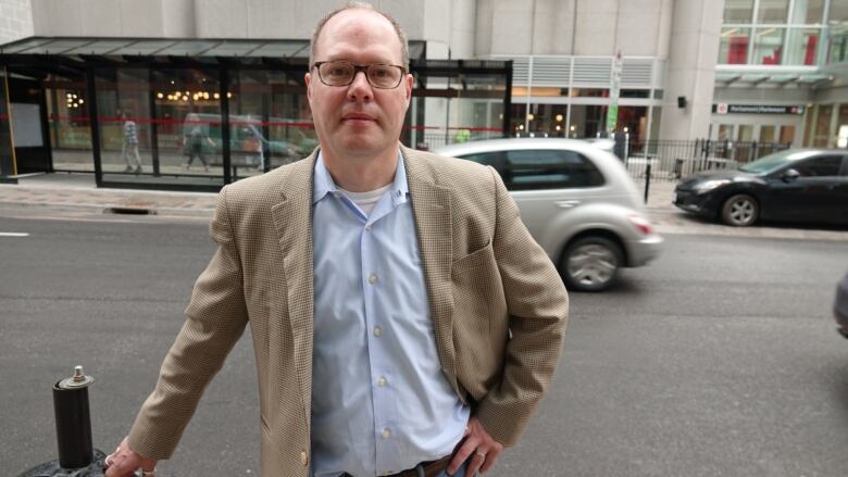 A man in a blazer and collared shirt stands on a downtown street as a car passes behind him.