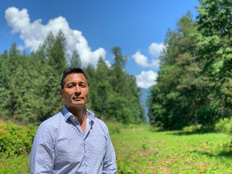 Man stands in a collared shirt in front of a lush green backdrop and blue skies.