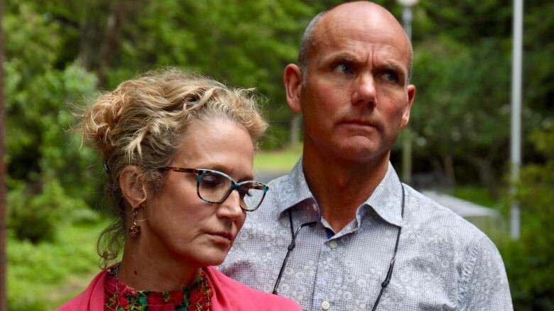 Two parents look sadly at the ground with foliage behind them.