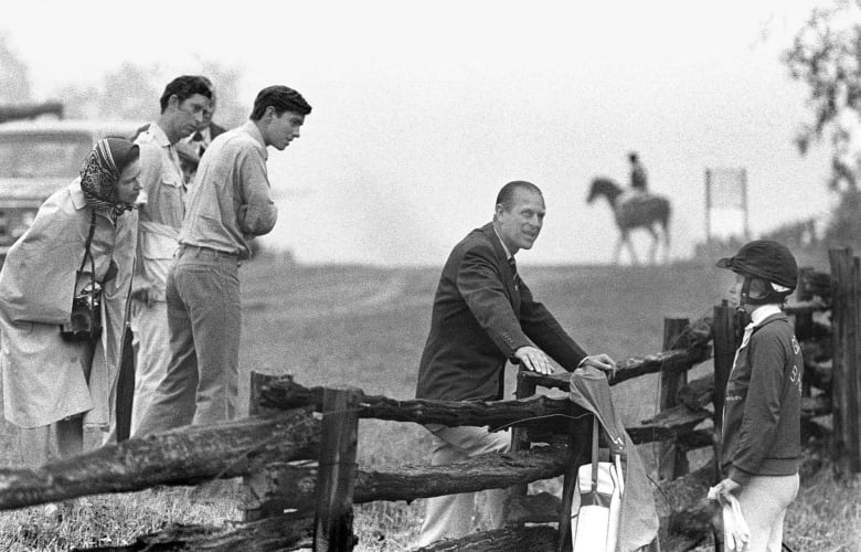 A person leans on a fence while talking with another person dressed in equestrian gear while other people look on.