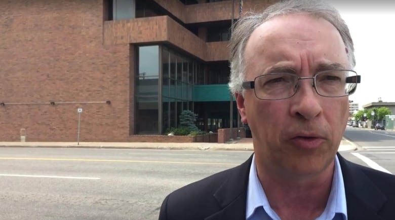 John Rustad gestures as he speaks in front of a building on a street. He's a white man with white hair and glasses.