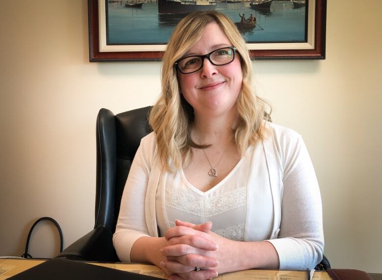 A woman with glasses, blonde hair and a white blouse and sweater sits at a desk and smiles.